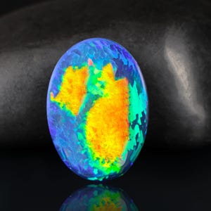 May include: Oval-shaped opal gemstone with a vibrant display of colors. The opal features a base of blue and green hues, with striking patches of orange and yellow. The gemstone is set against a dark background, highlighting its iridescent qualities.