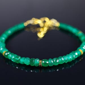 Puede incluir: Un brazalete de piedras preciosas verdes con detalles dorados. El brazalete está hecho de pequeñas cuentas facetadas y está enhebrado en una cadena delicada.
