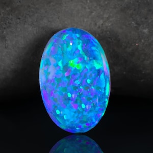 May include: An oval-shaped cabochon gemstone featuring a vibrant play-of-color with intense shades of electric blue, neon green, and deep violet. The surface displays a mosaic-like pattern of iridescent flashes that shift across the polished stone.