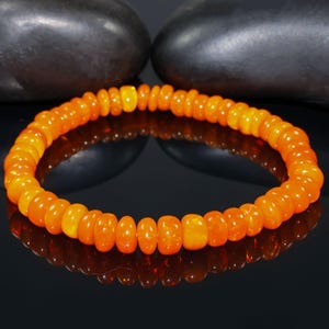 Puede incluir: Un vibrante brazalete de cuentas naranjas. Las cuentas son una mezcla de formas redondeadas y oblongas, con un acabado brillante. El brazalete se exhibe sobre una superficie negra reflectante, con piedras de color gris oscuro en el fondo.
