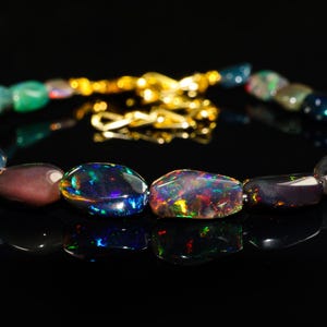 Puede incluir: Un collar hecho de piedras de ópalo negro iridiscente con un cierre de cadena de oro. Las piedras son de forma ovalada y tienen una variedad de colores, incluyendo verde, azul y morado.