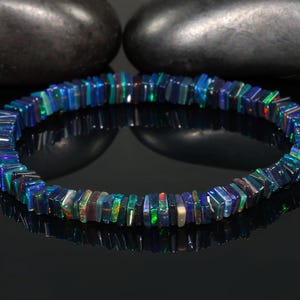 Könnte beinhalten: Ein Armband aus kleinen, rechteckigen, irisierenden blauen und grünen Opalsteinen. Die Steine sind zu einer kreisförmigen Form aufgereiht. Das Armband wird auf einer reflektierenden schwarzen Oberfläche mit zwei dunkelgrauen Steinen im Hintergrund präsentiert.