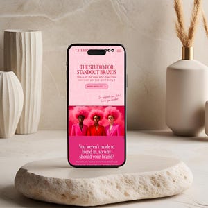Op de afbeelding: Een smartphone toont een website met de tekst "THE STUDIO FOR STANDOUT BRANDS". Het scherm toont drie vrouwen in roze pakken en pruiken. De telefoon ligt op een getextureerde, gebroken witte steen, met vazen op de achtergrond.