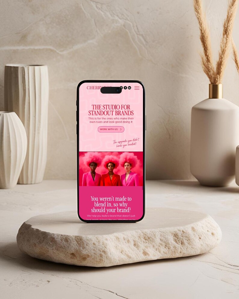 Op de afbeelding: Een smartphone toont een website met de tekst "THE STUDIO FOR STANDOUT BRANDS". Het scherm toont drie vrouwen in roze pakken en pruiken. De telefoon ligt op een getextureerde, gebroken witte steen, met vazen op de achtergrond.