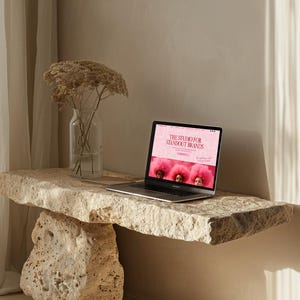 Op de afbeelding: Een laptop met de website "THE STUDIO FOR STANDOUT BRANDS" staat op een natuurstenen tafel. Links staat een heldere glazen vaas met gedroogde bloemen. De tafel wordt ondersteund door een grote stenen voet.