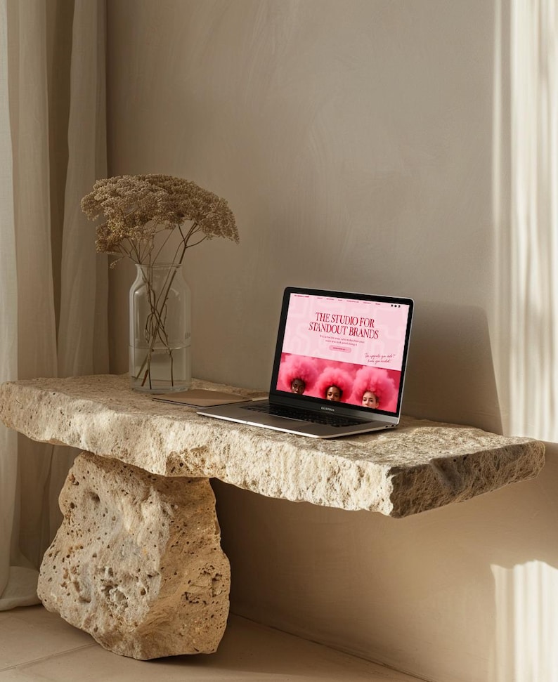 Op de afbeelding: Een laptop met de website "THE STUDIO FOR STANDOUT BRANDS" staat op een natuurstenen tafel. Links staat een heldere glazen vaas met gedroogde bloemen. De tafel wordt ondersteund door een grote stenen voet.