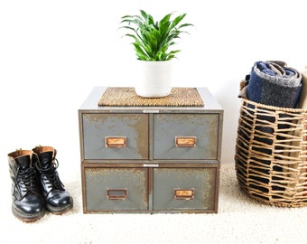 May include: A four-drawer metal cabinet with a woven mat on top, a potted green plant, and a wicker basket filled with folded blankets. Two black leather boots are on the floor in front of the cabinet.
