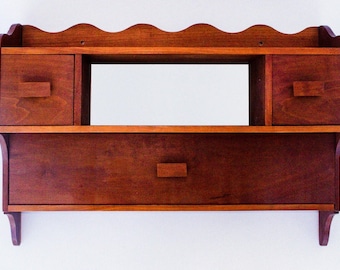 May include: A dark brown wooden wall shelf with three drawers and a shelf. The shelf has a scalloped edge and the drawers have simple wooden knobs.