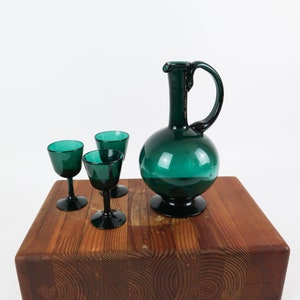 May include: A set of four teal green glass pieces, including a pitcher and three small glasses. The glasses have a rounded bowl and a stem. The pitcher has a round body and a curved handle. The pieces are arranged on a wooden surface.