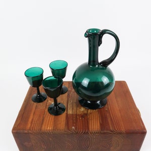 May include: A set of four emerald green glass decanter and shot glasses. The decanter has a round body and a long neck with a handle. The shot glasses are small and have a round bowl and a stem.