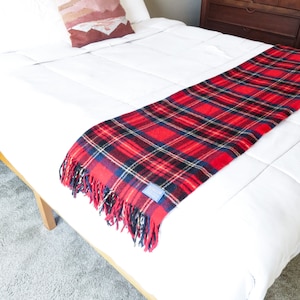 Puede incluir: Una manta de tiro de cuadros rojos y negros con bordes de flecos está colocada sobre una cama blanca. La manta tiene una etiqueta azul con texto blanco.