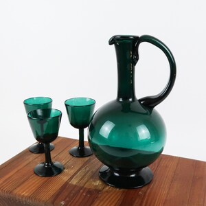 May include: A set of four emerald green glass decanter and glasses. The decanter has a round body and a long neck with a handle. The glasses are small and have a round bowl and a stem.