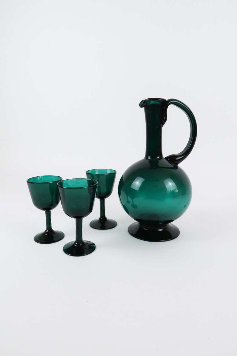 May include: A set of three teal green glass shot glasses and a matching teal green glass decanter with a round body and a curved handle.