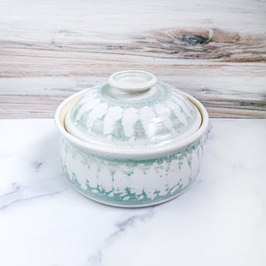 May include: A white ceramic bowl with a lid, featuring a light blue and white speckled glaze. The bowl has a rounded shape and a slightly flared rim.