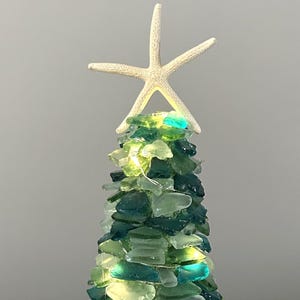 May include: A decorative tree made of sea glass in shades of green and blue, illuminated with small lights. A white starfish tops the tree. The tree is on a wooden surface.