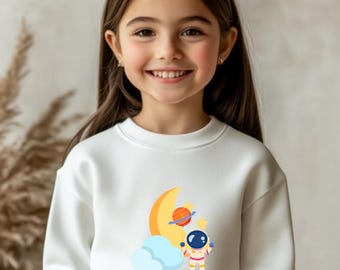 May include: White sweatshirt featuring a colorful graphic of an astronaut, a crescent moon, a planet, and a cloud. The design is in a cartoon style. The sweatshirt is worn by a young person.