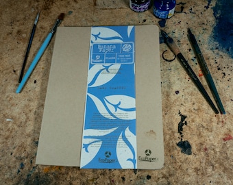 Könnte beinhalten: Ein rechteckiges, leeres Notizbuch aus Bananenpapier mit einem blau-weißen Etikett mit der Aufschrift "Banana Paper" und "Hand Crafted". Zwei Pinsel und zwei Kalligraphie-Stifte liegen auf der Holzoberfläche.