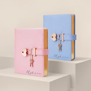 May include: Two journals, one pink and one blue, with gold-edged pages. Each journal has a heart-shaped lock with a key charm and a strap closure. The word "Ushine" is written on the bottom of each journal.