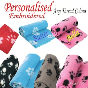 May include: Six personalised fleece blankets with paw print designs. The blankets are rolled up and feature embroidered names, including "Buster" and "Bella".