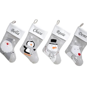 May include: Four grey Christmas stockings with white cuffs and hanging loops. Each stocking features a different festive character: Santa Claus, a penguin, a snowman, and a reindeer. The names "Bella", "Chase", "Rosie", and "Demi" are embroidered on the cuffs.