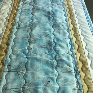 May include: A blue quilted table runner with a wintery forest scene featuring bare trees and small birds. The runner has a scalloped edge and is trimmed with gold and white fabric.