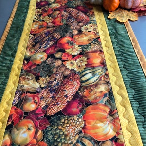 May include: A quilted table runner with a fall harvest theme. The runner features a dark background with a variety of fruits and vegetables, including apples, pumpkins, gourds, and corn. The runner has a green and yellow border.