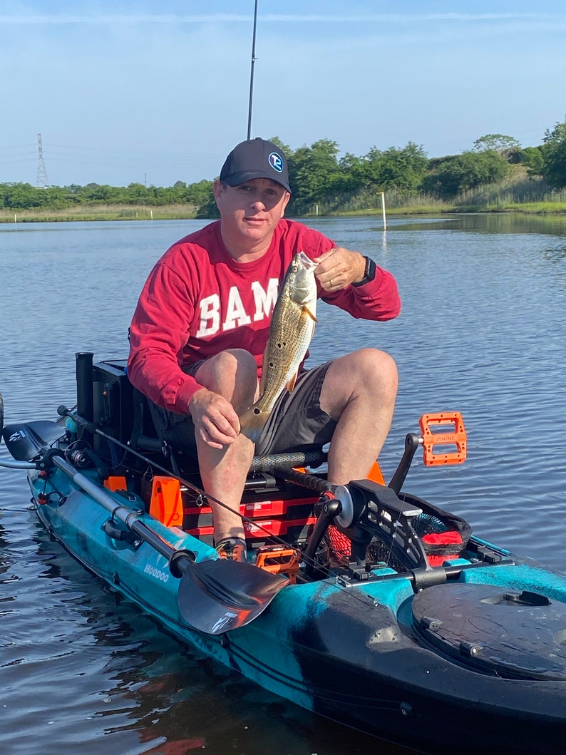 May include: A person wearing a red long-sleeve shirt with the word "BAM" printed on it, a black baseball cap, and black shorts, is sitting in a blue and black kayak. The person is holding a red fish with their right hand. The kayak has a black paddle and orange and black accessories.