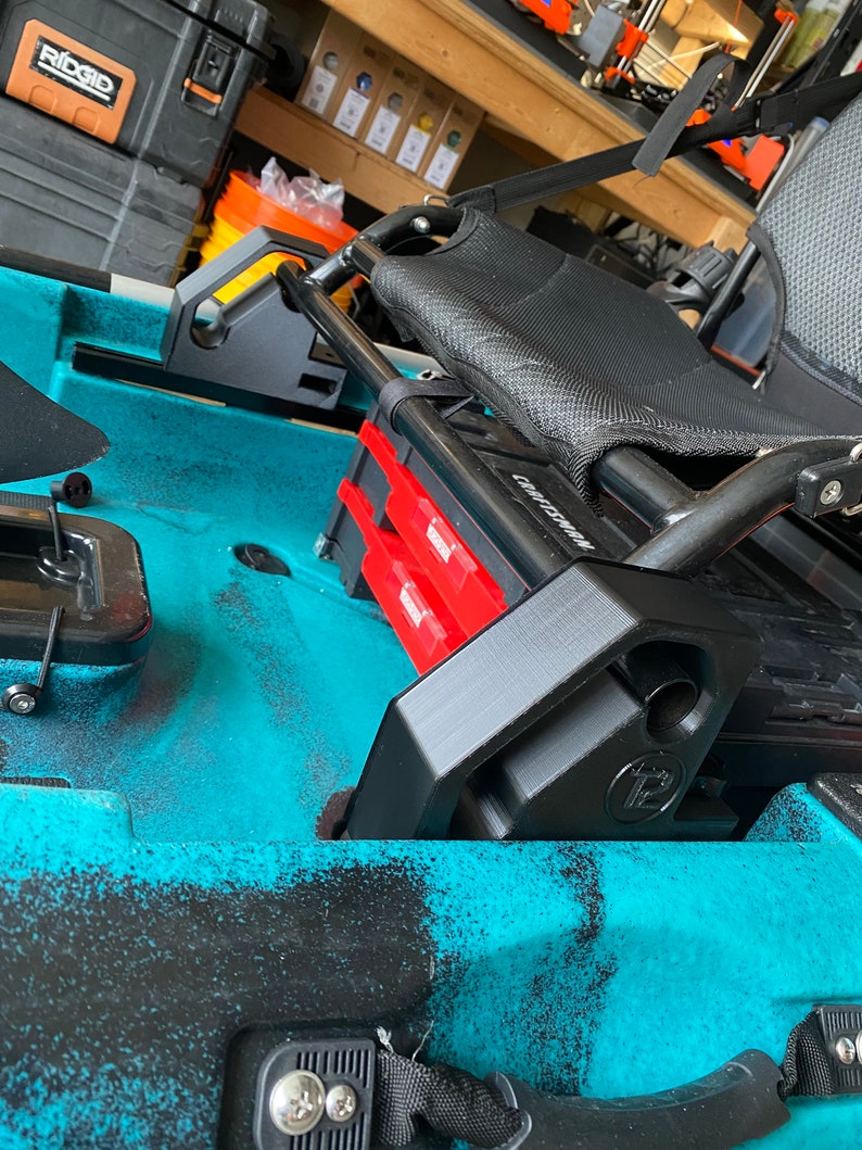 May include: A black kayak seat with a black mesh backrest and a black metal frame. The seat is mounted on a blue kayak with a black storage box and a red tackle box.