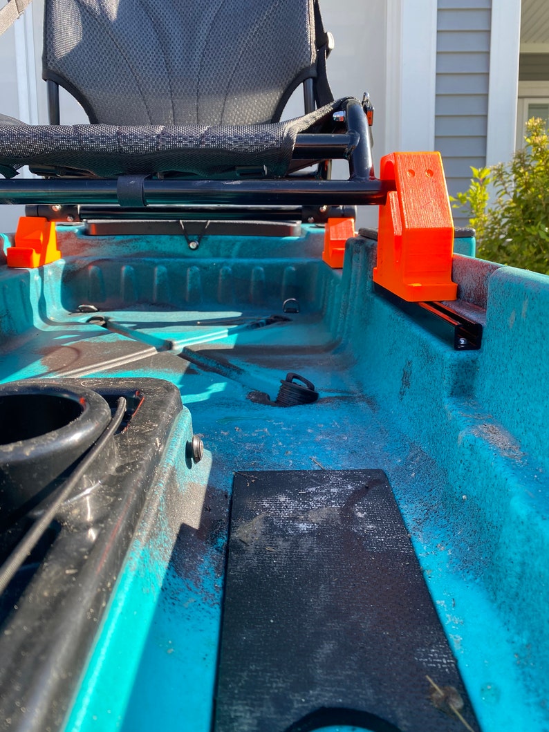 May include: A teal kayak with a black seat and orange plastic supports. The seat is raised above the kayak deck.
