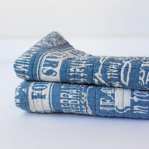 May include: Two folded blue and white quilts with a printed design. The quilts are stacked on top of each other, showcasing the detailed pattern and texture of the fabric. The design includes text and decorative elements.