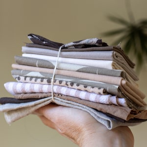 May include: A stack of folded fabric squares in various neutral colors, including beige, brown, and white, tied together with twine. The fabrics have different patterns, such as stripes and polka dots.