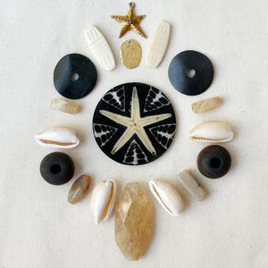May include: A collection of assorted beads and charms arranged in a circular pattern, including a large black circular disc with a white starfish design, white cowrie shells, dark wooden beads, smooth tan stones, and a gold-toned starfish charm.