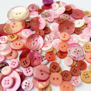 May include: A collection of assorted sewing buttons in various shades of pink, red, orange, and cream. The buttons vary in size, texture, and material, featuring two or four holes for attachment. Some have a glossy finish, while others are matte.