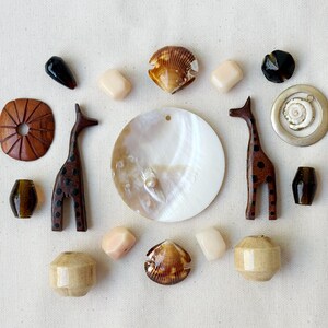 May include: A collection of craft beads and pendants including two wooden giraffe figures, a large circular mother-of-pearl shell, two brown seashell halves, a silver-toned metal ring with a small shell charm, and various smooth stone and wooden beads.