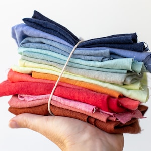 May include: A stack of folded fabric squares in a spectrum of colors including dark blue, light blue, teal, yellow, orange, red, pink, and brown. The fabric pieces are held together by a thin, light-colored string wrapped around the center.