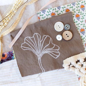 May include: A collection of craft supplies including fabric scraps with floral and leaf patterns, various buttons, and decorative ribbons. A piece of brown fabric features a white embroidered leaf design. The items are arranged on a light-colored surface.