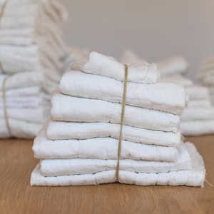 May include: A stack of folded white linen cloths, secured with a natural twine. The cloths have a quilted texture and are neatly arranged on a wooden surface. Additional stacks of similar cloths are visible in the background, suggesting a set or collection.