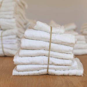 May include: A stack of white rectangular fabric cloths secured with a thin tan rubber band. The cloths feature a quilted texture with visible stitching lines running horizontally across the surface, creating a layered and soft appearance.