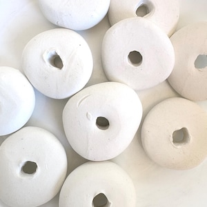 May include: A collection of white, donut-shaped ceramic beads with a central hole. The beads have a slightly textured surface and vary in size and shape. The arrangement is on a white surface, creating a clean, minimalist aesthetic.