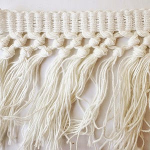 May include: Close-up of a cream-colored macrame fringe trim. The trim features a woven top section, knotted details, and long, flowing fringe. This decorative trim is suitable for various craft and design projects.