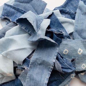 May include: A pile of various denim fabric scraps in shades of blue. The pieces vary in size and shape, with some featuring a diamond pattern. The edges are frayed, suggesting a repurposed or upcycled craft project.