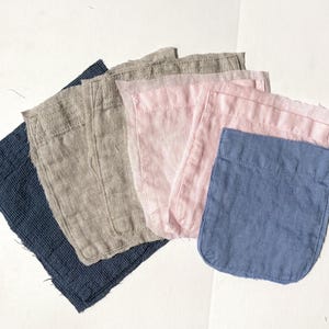 May include: A collection of linen fabric pockets in various colors, including navy blue, tan, light pink, and a lighter blue. The pockets are rectangular with a curved bottom edge and are arranged on a white surface.