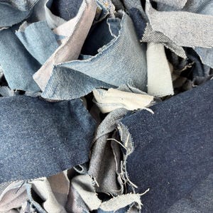 May include: A collection of assorted denim fabric scraps in various shades of blue, ranging from light wash to dark indigo. The pieces feature frayed, uneven edges and visible twill weave textures, layered together in a dense, overlapping pile.