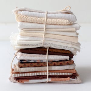 May include: A stack of assorted fabric scraps in various shades of white, cream, brown, and orange, tied together with twine. The fabrics include plain weaves, lace, and patterned textiles, creating a rustic, vintage aesthetic. Ideal for quilting or crafting projects.