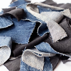 May include: A pile of denim fabric scraps in various shades of blue and black. The pieces are irregularly shaped with frayed edges, suggesting a craft or upcycling project. The textures and colors create a visually interesting composition.