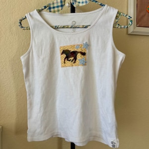 May include: A white sleeveless tank top featuring a rectangular patch on the chest. The patch displays a dark horse silhouette against a yellow background with light blue star-shaped accents. A small floral detail is visible at the bottom hem.