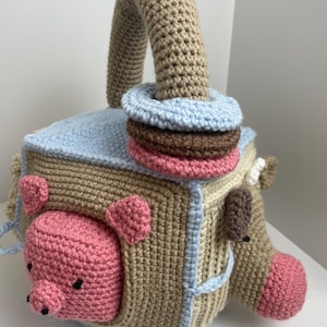 May include: A handmade crochet toy cube with a tan base, light blue accents, and a tan handle. The cube features a pink pig, a brown horse, and a stack of colorful rings. This soft toy is designed for babies and toddlers.