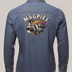 May include: A denim shirt with a Magpies logo embroidered on the back. The logo shows a cartoon magpie in black, white, and yellow, holding a football. The shirt has long sleeves and a classic collar. The shirt is made of denim.