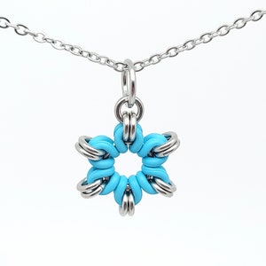 May include: A silver-toned necklace with a star-shaped pendant. The pendant features a bright turquoise center surrounded by silver metal rings, creating a flower-like design. The chain is delicate and hangs against a white background.