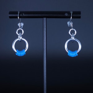 May include: A pair of silver-toned hoop earrings with a bright blue accent. Each earring features a large circular hoop with a series of small blue rings at the bottom. The earrings hang from a simple hook.